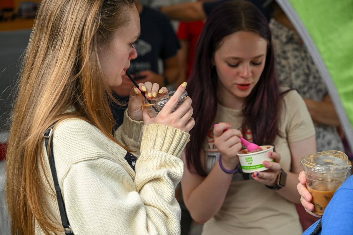 Two customers standing under the tent at the soft opening, one drinking an iced coffee through a straw and the other eating a cup of ice cream.