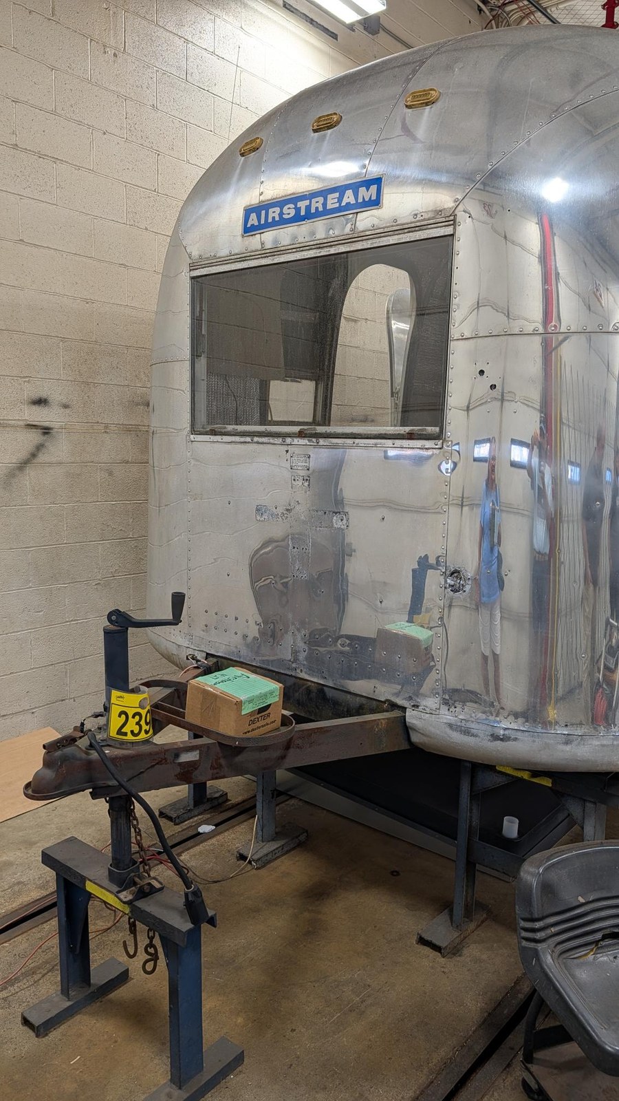 The front of the Airstream raised on stands, with the trailer tongue and coupler supported by a jack and a boxed Dexter axle component resting on the frame rail.