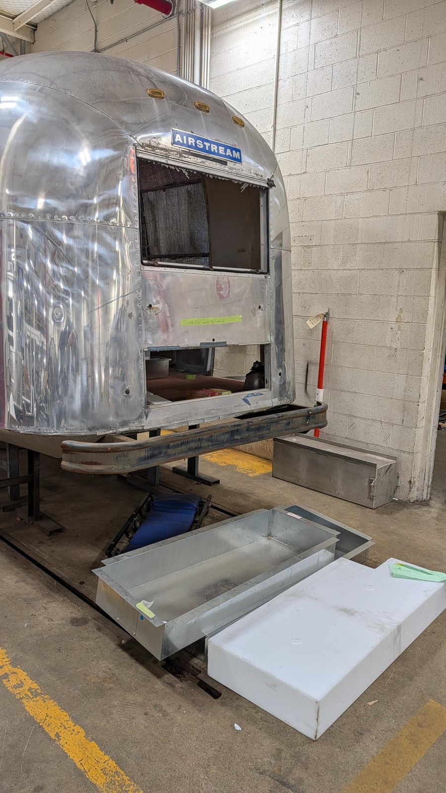 The Airstream's front end with a window and lower side panel removed, and new galvanized sheet metal ducting and a white holding tank staged on the shop floor.