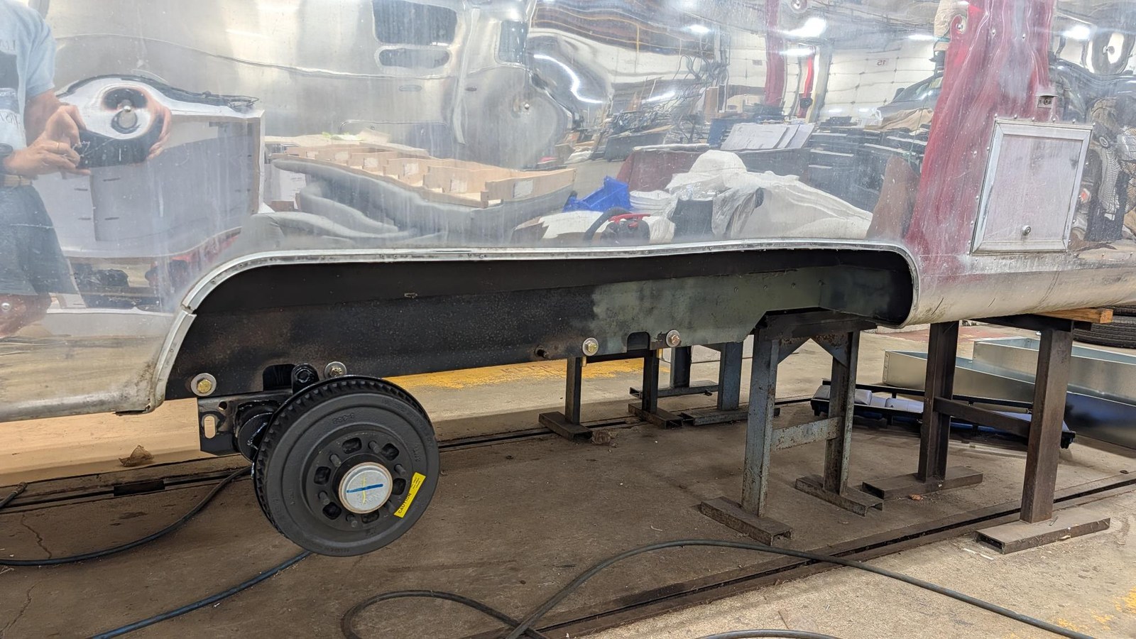 The underside of the Airstream with the belly pan opened up, showing the frame resting on stands and a newly fitted axle hub and wheel.