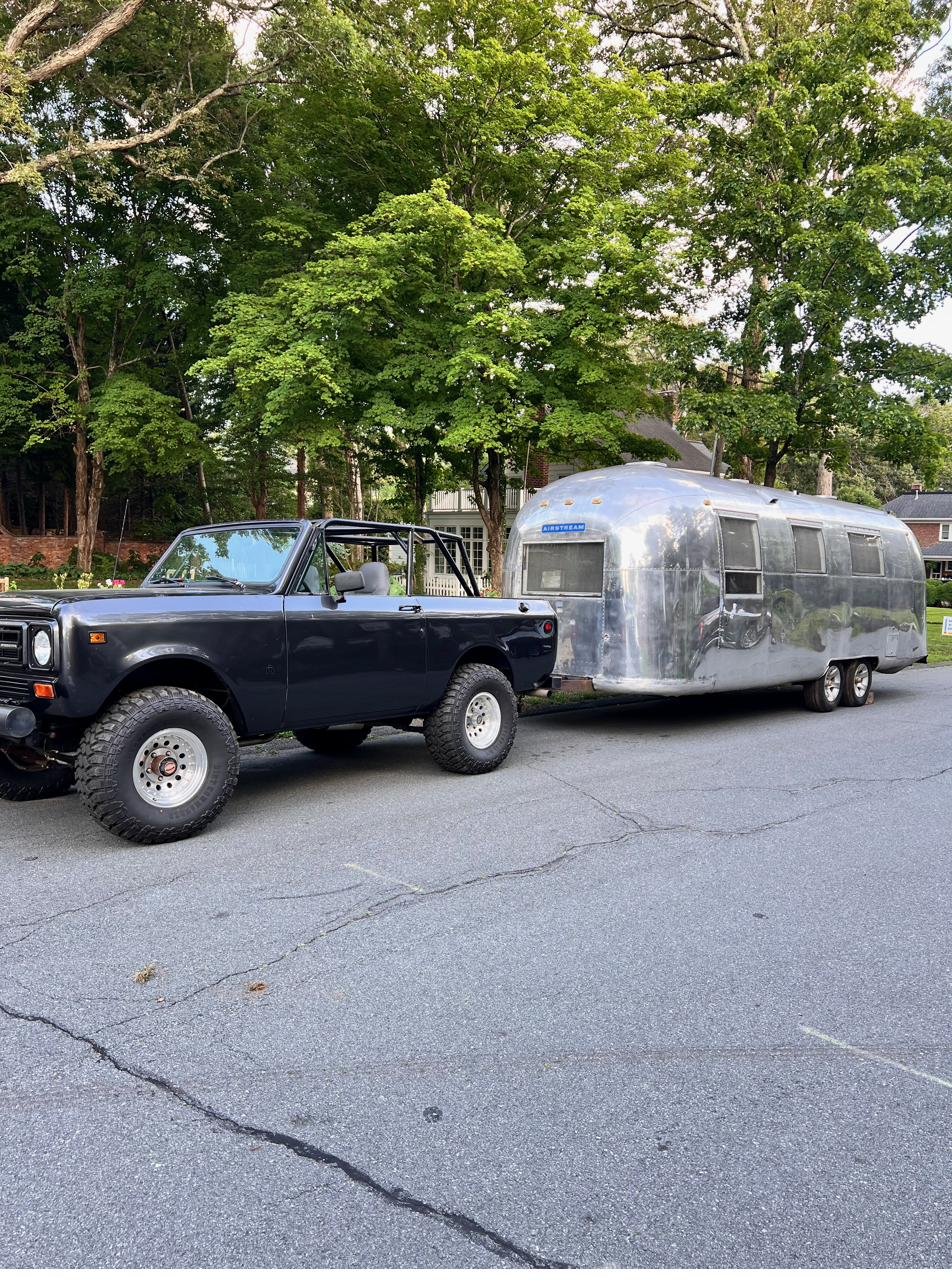 The black 1979 International Scout II hitched to the polished Silver Bullet Airstream, both parked on a tree-lined street.