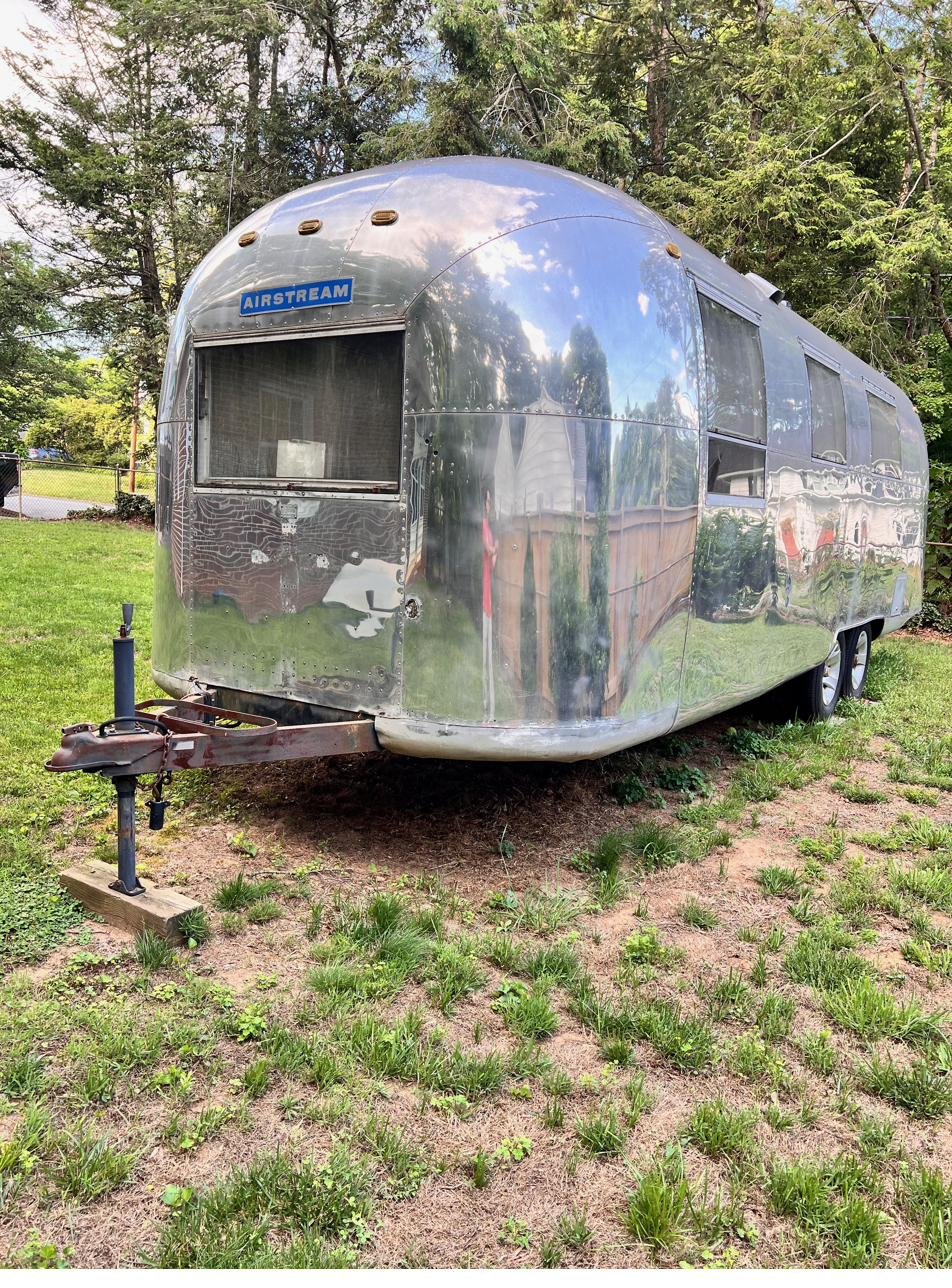 Front three-quarter view of the polished Silver Bullet Airstream, the AIRSTREAM badge on the nose and trees mirrored in the riveted aluminum skin.